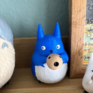 May include: A blue figurine of a cartoon character with white belly and holding a small, round object. The figurine has large eyes and pointed ears. The object it holds is light brown with a dark hole.