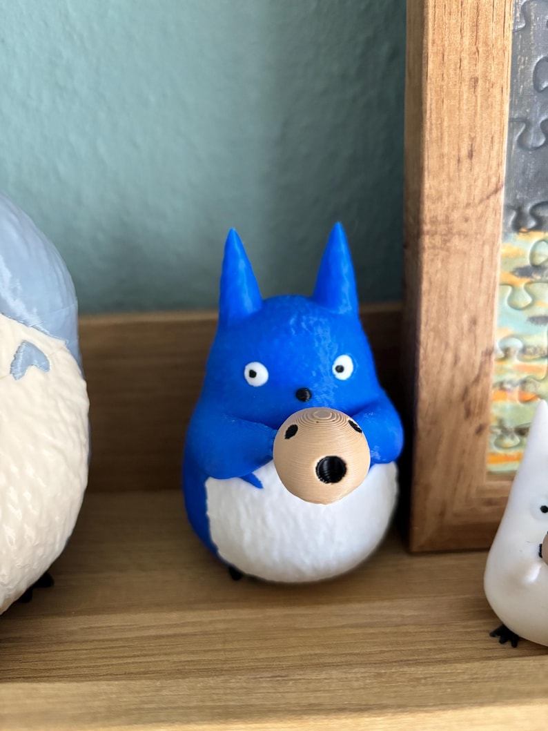 May include: A blue figurine of a cartoon character with white belly and holding a small, round object. The figurine has large eyes and pointed ears. The object it holds is light brown with a dark hole.