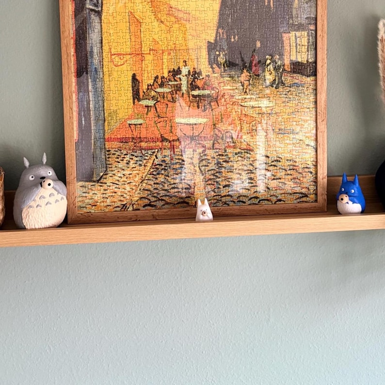 May include: A wooden framed jigsaw puzzle depicting a cafe scene, displayed on a wooden shelf with three small, decorative figurines. The figurines are in the shape of cartoon characters, one gray, one white, and one blue.