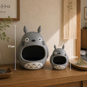 Può includere: Due contenitori grigi a forma di Totoro, uno alto 17 cm e l'altro 10 cm, sono su una superficie di legno. I contenitori hanno grandi aperture e dettagli bianchi sulla pancia. Lo sfondo include una stampa incorniciata e un vaso.