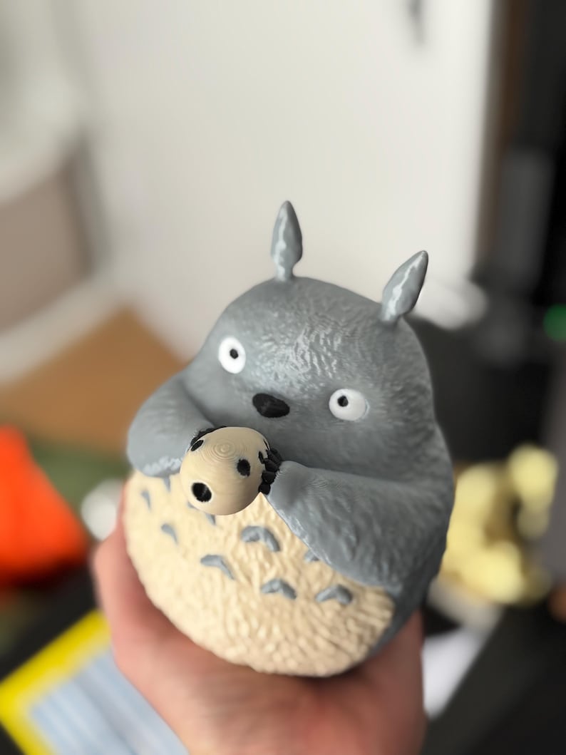 May include: A gray Totoro figurine holding a small, round, light-colored object with black spots. The figurine has large, white eyes and small, pointed ears. The bottom portion is a light beige color with small, dark gray markings.