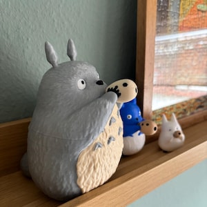 May include: A gray Totoro figurine holding a small, round object, with a cream-colored belly. Beside it are two smaller figurines: a blue one and a white one. All are displayed on a wooden shelf against a light blue wall.