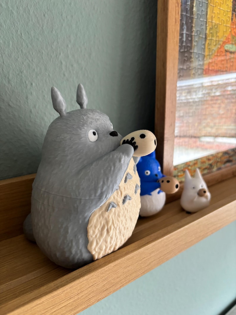May include: A gray Totoro figurine holding a small, round object, with a cream-colored belly. Beside it are two smaller figurines: a blue one and a white one. All are displayed on a wooden shelf against a light blue wall.