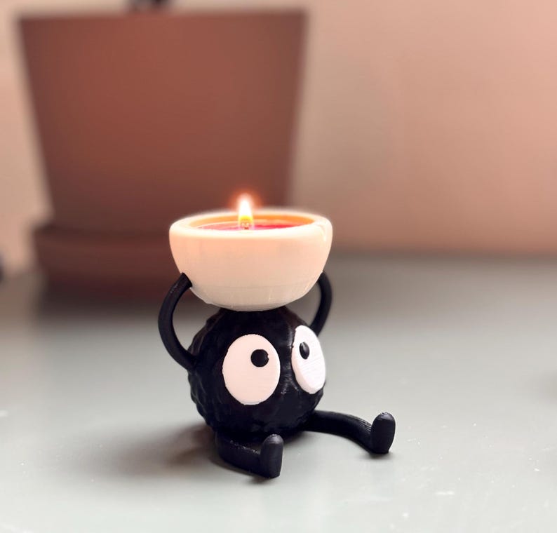Adorable 3D-printed Soot Sprite tealight holder inspired by Studio Ghibli. This black and white candle holder features a cute soot spirit figure holding a small tealight, creating a cozy, magical ambiance. Perfect for anime fans, home decor lovers, and unique gift ideas. Add a touch of whimsy to your space with this handcrafted, geeky candle holder. Great for desks, shelves, or nightstands!