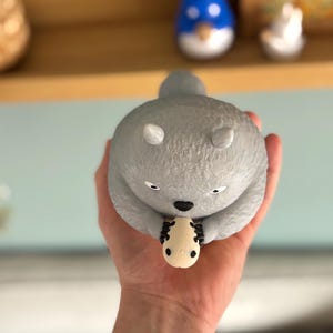 May include: A gray, round figurine of a koala holding a small, cream-colored object. The koala has white ears and black eyes. A blue and white figurine is visible in the background.