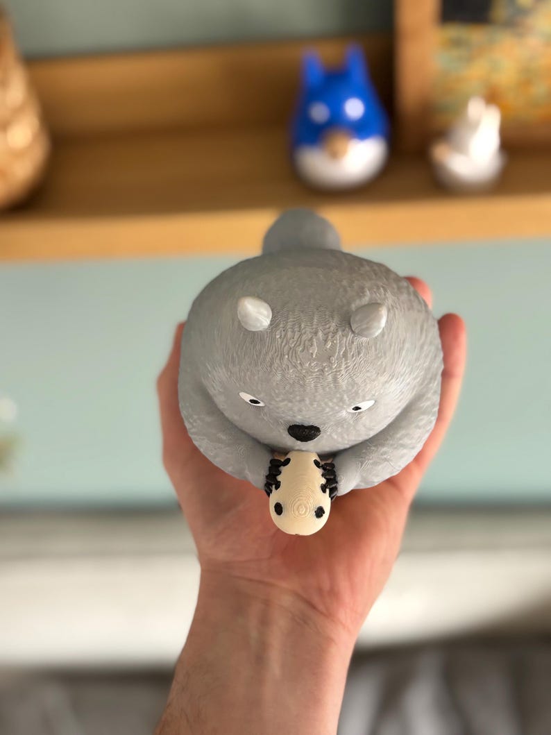 May include: A gray, round figurine of a koala holding a small, cream-colored object. The koala has white ears and black eyes. A blue and white figurine is visible in the background.