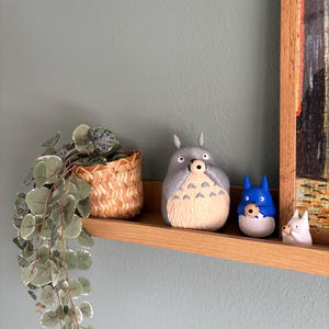 May include: A wooden shelf displays three cartoon figurines: a large gray Totoro, a blue character, and a small white figure. A woven basket with a trailing plant is to the left. The shelf is against a light blue wall.