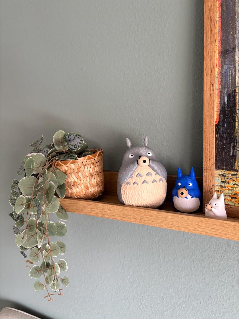 May include: A wooden shelf displays three cartoon figurines: a large gray Totoro, a blue character, and a small white figure. A woven basket with a trailing plant is to the left. The shelf is against a light blue wall.