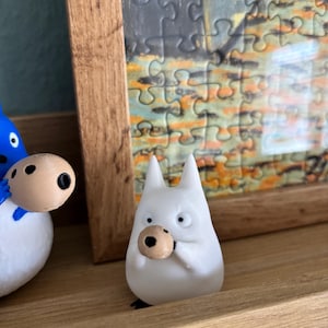 May include: A white figurine of a cartoon character with pointed ears and a round, tan object held in its hands. The figurine is on a wooden shelf, with a framed puzzle in the background.