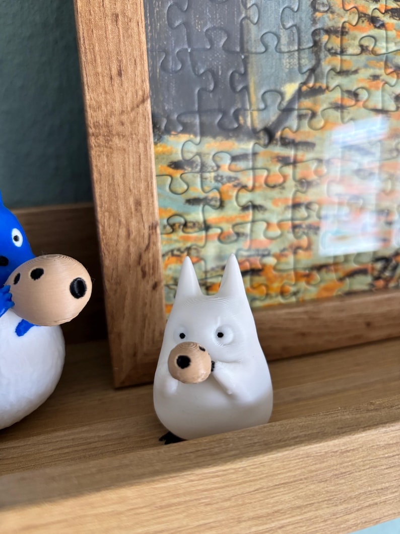May include: A white figurine of a cartoon character with pointed ears and a round, tan object held in its hands. The figurine is on a wooden shelf, with a framed puzzle in the background.