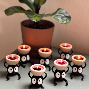 Adorable 3D-printed Soot Sprite tealight holder inspired by Studio Ghibli. This black and white candle holder features a cute soot spirit figure holding a small tealight, creating a cozy, magical ambiance. Perfect for anime fans, home decor lovers, and unique gift ideas. Add a touch of whimsy to your space with this handcrafted, geeky candle holder. Great for desks, shelves, or nightstands!