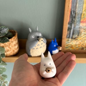 May include: A white figurine of a Totoro-like character with a small brown object in its mouth, held in a hand. Other figurines of similar characters are on a shelf in the background, including a gray and a blue one.