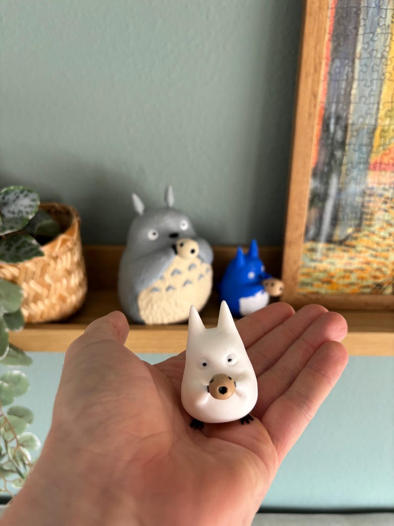 May include: A white figurine of a Totoro-like character with a small brown object in its mouth, held in a hand. Other figurines of similar characters are on a shelf in the background, including a gray and a blue one.