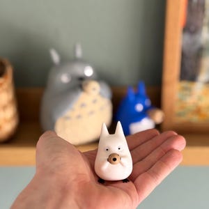 May include: A small, white figurine of a cartoon character with pointed ears and a round, brown object held in its hands. The figurine is held in a person's hand, with other similar figurines visible in the background.