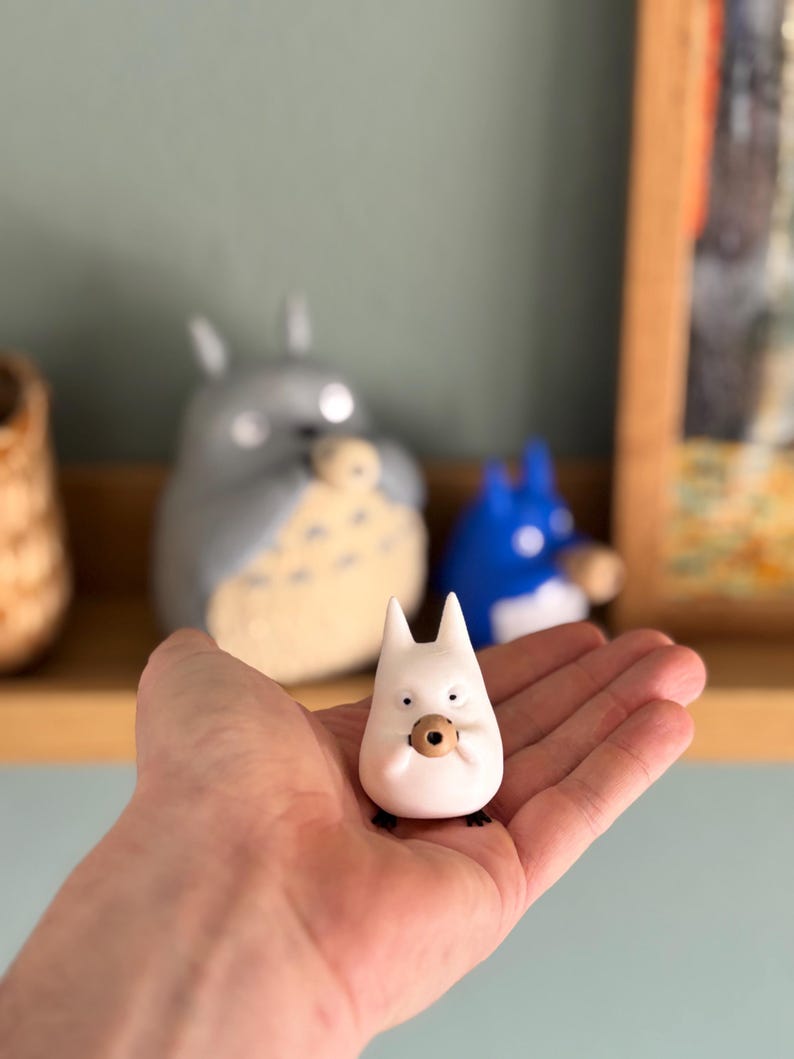 May include: A small, white figurine of a cartoon character with pointed ears and a round, brown object held in its hands. The figurine is held in a person's hand, with other similar figurines visible in the background.