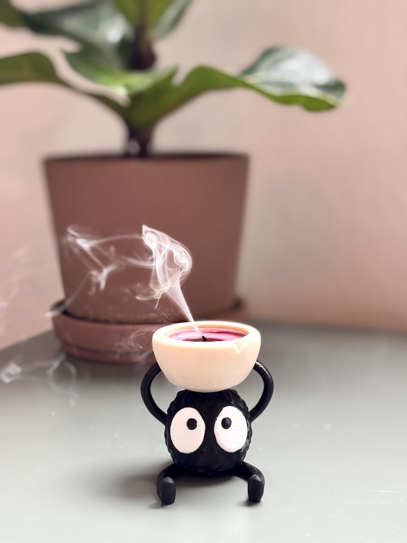 Adorable 3D-printed Soot Sprite tealight holder inspired by Studio Ghibli. This black and white candle holder features a cute soot spirit figure holding a small tealight, creating a cozy, magical ambiance. Perfect for anime fans, home decor lovers, and unique gift ideas. Add a touch of whimsy to your space with this handcrafted, geeky candle holder. Great for desks, shelves, or nightstands!