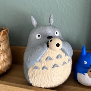May include: A gray Totoro figurine holding a small, round object. The figurine has a cream-colored belly with dark gray markings. It has large, round eyes and pointed ears. A blue Totoro figurine is visible to the right.