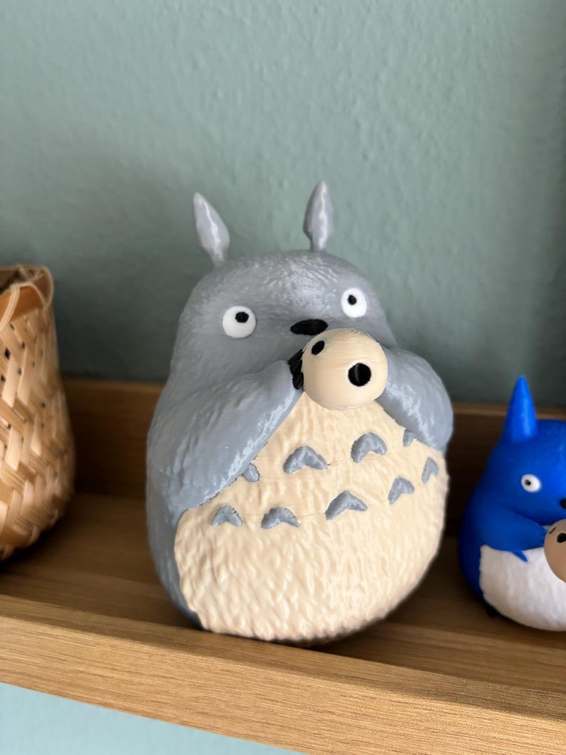 May include: A gray Totoro figurine holding a small, round object. The figurine has a cream-colored belly with dark gray markings. It has large, round eyes and pointed ears. A blue Totoro figurine is visible to the right.