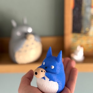 May include: A blue figurine of a cartoon character with a white belly and a tan nose. The figurine is held in a hand. Another similar figurine is in the background, along with a small white figurine.