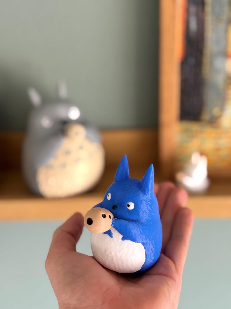 May include: A blue figurine of a cartoon character with a white belly and a tan nose. The figurine is held in a hand. Another similar figurine is in the background, along with a small white figurine.