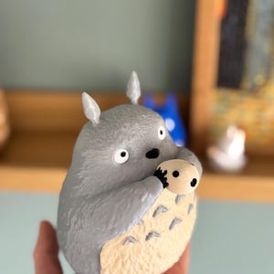 May include: A gray figurine of Totoro, a character from the animated film My Neighbor Totoro, holding a smaller, tan-colored creature. The figurine has large eyes, pointed ears, and a cream-colored belly. The figure is held in a hand.