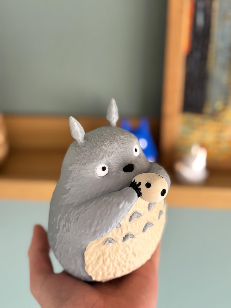 May include: A gray figurine of Totoro, a character from the animated film My Neighbor Totoro, holding a smaller, tan-colored creature. The figurine has large eyes, pointed ears, and a cream-colored belly. The figure is held in a hand.