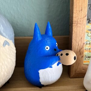May include: A blue and white figurine of a cartoon character holding a small, round object with black spots. The figurine has pointed ears and a white belly. The background includes a wooden frame and other figurines.