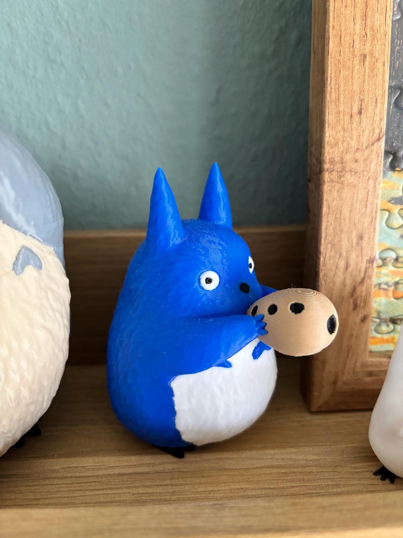 May include: A blue and white figurine of a cartoon character holding a small, round object with black spots. The figurine has pointed ears and a white belly. The background includes a wooden frame and other figurines.