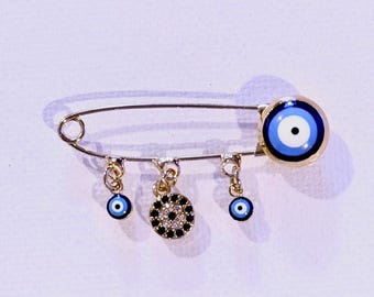 Puede incluir: Un imperdible dorado con colgantes. El imperdible presenta un gran dije de ojo azul y blanco en el extremo y tres dijes más pequeños. Un dije es un diseño circular en blanco y negro, y los otros dos son dijes de ojo azul y blanco.