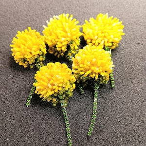 May include: Five handmade beaded dandelions with bright yellow flower heads and green stems. The flower heads are made of small yellow beads, and the stems are made of green beads. The dandelions are arranged on a dark gray surface.