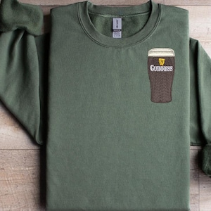 Puede incluir: Una sudadera verde con un logotipo de Guinness bordado en blanco en el pecho izquierdo. El logotipo presenta un vaso de cerveza con un arpa y la palabra "Guinness" debajo.