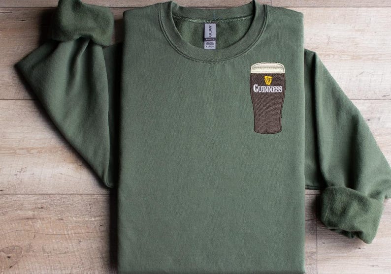 Puede incluir: Una sudadera verde con un logotipo de Guinness bordado en blanco en el pecho izquierdo. El logotipo presenta un vaso de cerveza con un arpa y la palabra "Guinness" debajo.
