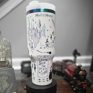May include: A white travel mug with a black base, featuring a detailed illustration of a castle, stars, and other graphics. The text "Mischief Managed" and "I Solemnly Swear That I'm Up to No Good" are also visible.