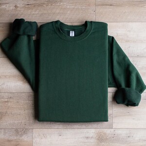Puede incluir: Una sudadera de cuello redondo de color verde oscuro con mangas largas. La sudadera est&aacute; colocada sobre una superficie de madera.
