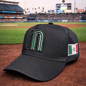 May include: Black baseball cap featuring a green and black embroidered "M" logo and a Mexican flag patch. The cap is set against a baseball field backdrop, with stadium lights and a crowd visible in the background.