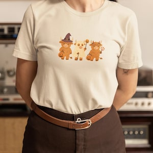 May include: A light beige t-shirt featuring a Halloween-themed graphic of three cartoon Highland cows. The cows are dressed in costumes, including a witch hat, holding a pumpkin, and a trident. The person is wearing a brown belt and brown pants.