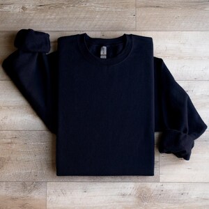 Puede incluir: Una sudadera negra de cuello redondo y mangas largas. La sudadera est&aacute; doblada y colocada sobre una superficie de madera.