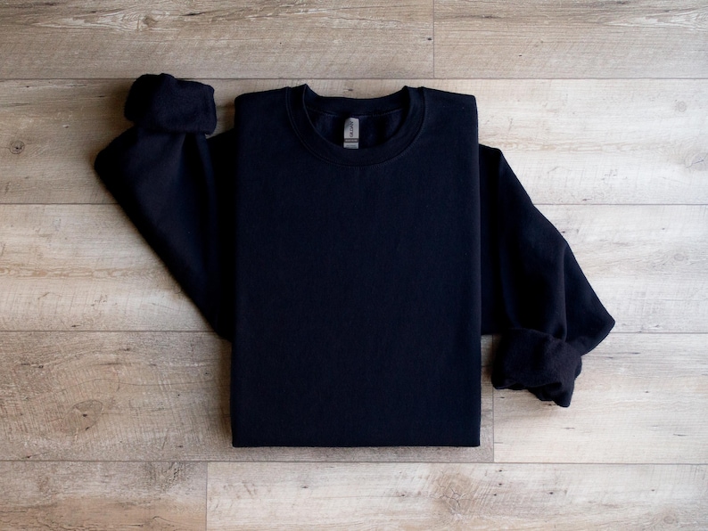 Puede incluir: Una sudadera negra de cuello redondo y mangas largas. La sudadera est&aacute; doblada y colocada sobre una superficie de madera.