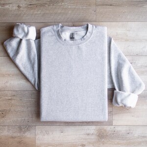 Puede incluir: Una sudadera gris lisa de manga larga y cuello redondo. La sudadera est&aacute; colocada sobre una superficie de madera.