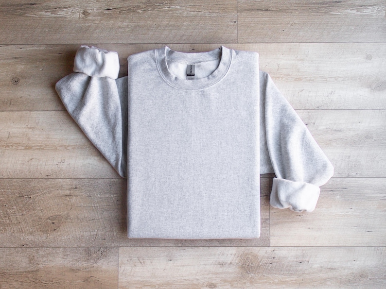 Puede incluir: Una sudadera gris lisa de manga larga y cuello redondo. La sudadera est&aacute; colocada sobre una superficie de madera.