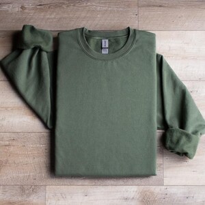 Puede incluir: Una sudadera verde de cuello redondo con mangas largas. La sudadera est&aacute; doblada por la mitad y colocada sobre una superficie de madera.