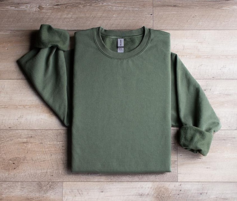Puede incluir: Una sudadera verde de cuello redondo con mangas largas. La sudadera est&aacute; doblada por la mitad y colocada sobre una superficie de madera.