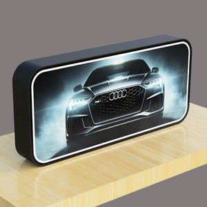 Peut inclure: Haut-parleur rectangulaire noir avec une bordure blanche, affichant une image d'une Audi RS argentée. Les phares de la voiture sont allumés, et le logo Audi est visible. Le haut-parleur est posé sur une surface en bois.