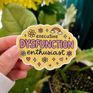 May include: A yellow sticker with the words "executive DYSFUNCTION enthusiast" in pink and yellow. The sticker is decorated with flower, heart, and diamond graphics, and a rainbow. The sticker is held in a hand.