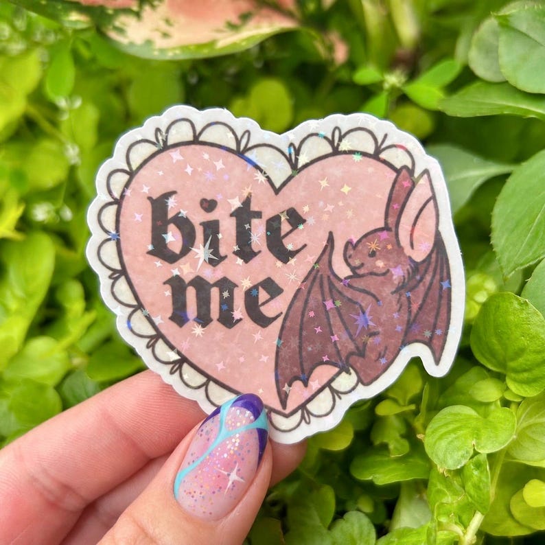 Hand holding a holographic “Bite Me” bat vinyl sticker featuring a pink heart, gothic black lettering, and an adorable brown bat, with rainbow holographic stars shimmering in natural sunlight against lush green plants.
