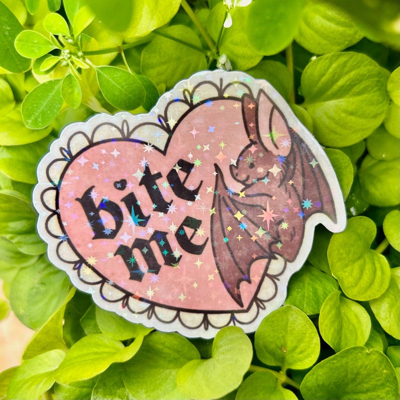 Close-up of the holographic “Bite Me” bat die-cut sticker resting among bright green leaves, highlighting the pastel pink heart, gothic typography, cute bat illustration, and rainbow holographic star laminate reflecting in the light.