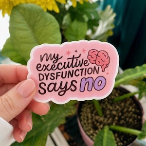 May include: A pink sticker with the text "my executive dysfunction says no" in black and purple. The sticker has a brain graphic and is held by a person's hand in front of a plant.