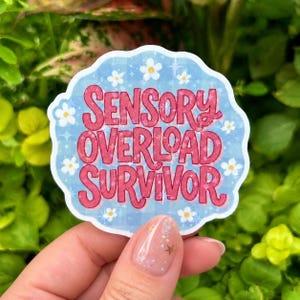 Hand holding a glossy holographic vinyl sticker reading “Sensory Overload Survivor” in bold pink retro lettering on a soft blue background with white daisies and sparkling stars. Photographed outdoors against lush green foliage with a sheer glitter manicure featuring gold stars and iridescent sparkle accents.