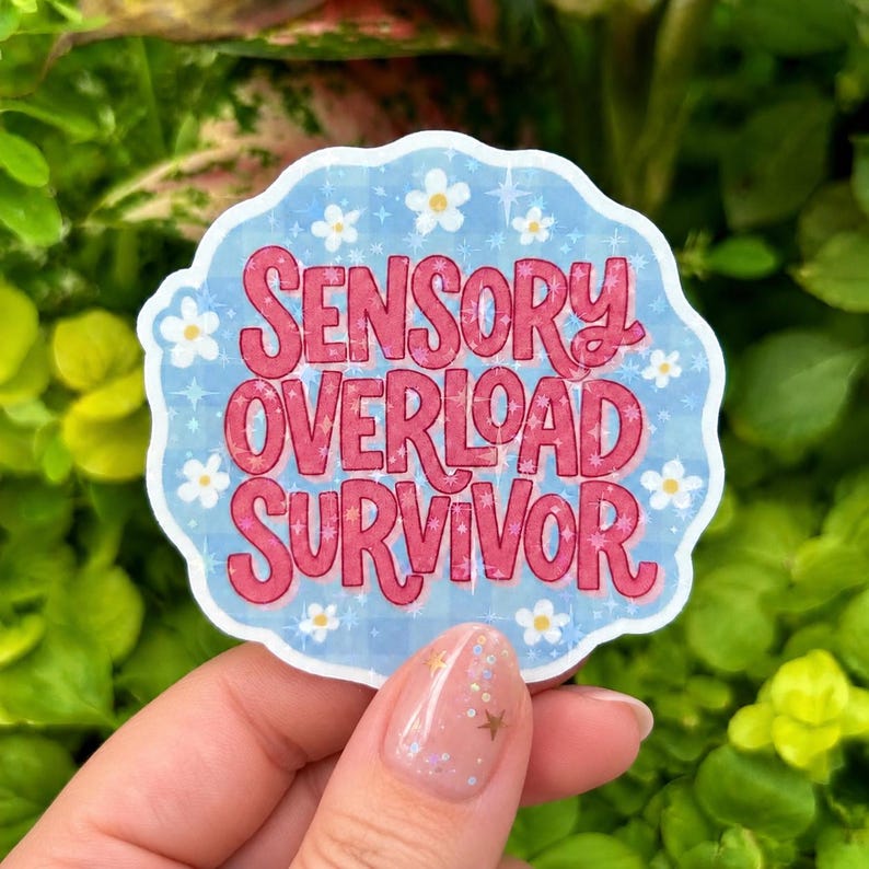 Hand holding a glossy holographic vinyl sticker reading “Sensory Overload Survivor” in bold pink retro lettering on a soft blue background with white daisies and sparkling stars. Photographed outdoors against lush green foliage with a sheer glitter manicure featuring gold stars and iridescent sparkle accents.