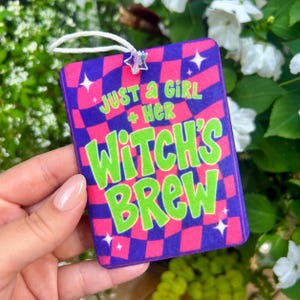 May include: A rectangular air freshener featuring a purple and pink checkered pattern with the text "JUST A GIRL + HER WITCH'S BREW" in bright green letters. The design includes small white star accents and a white hanging string with a star bead.